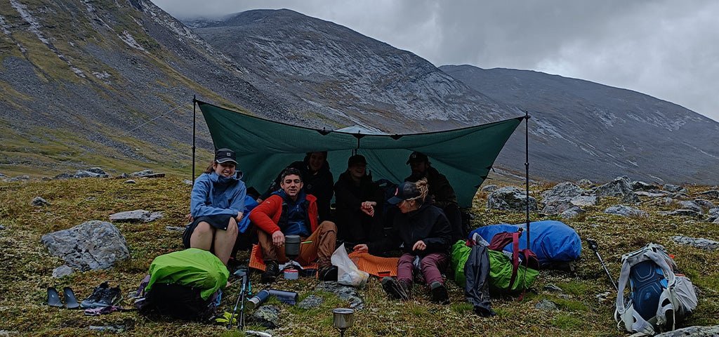 Group of campers under a tarp in a mountainous area, equipped with Fjallraven clothing and camping gear.