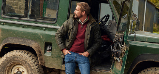 A man wearing a green Barbour jacket and red jumper stands confidently beside an open door of a rugged green Land Rover, set against a rural backdrop.