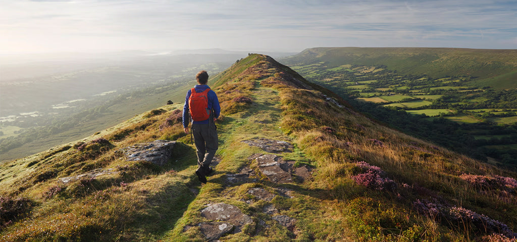 Man hiking on a rocky trail wearing a backpack and outdoor clothing, overlooking a scenic green valley at sunrise.