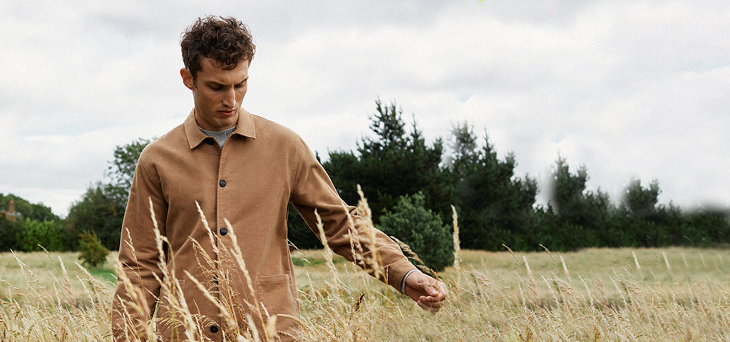 A man in a Sunspel shirt walking thoughtfully through a field with tall grass and trees in the background.