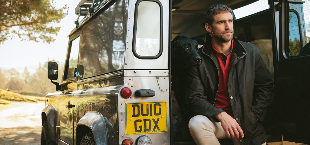 Barbour jacket showcased by a man leaning against a Land Rover with his dog. Durable and stylish outdoor wear ideal for countryside adventures and rugged terrains.