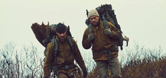 Filson Tin Cloth Cruiser jackets worn by two hunters trekking through rugged terrain with heavy packs in wet conditions