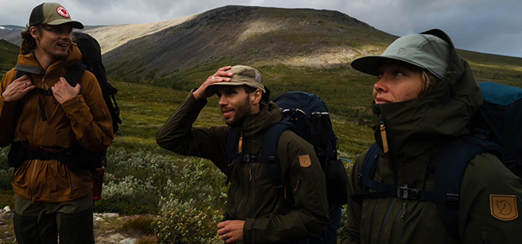 Hikers wearing Fjallraven outdoor clothing and backpacks pause on a trail in rugged terrain, ready for the Fjallraven Classic trek.