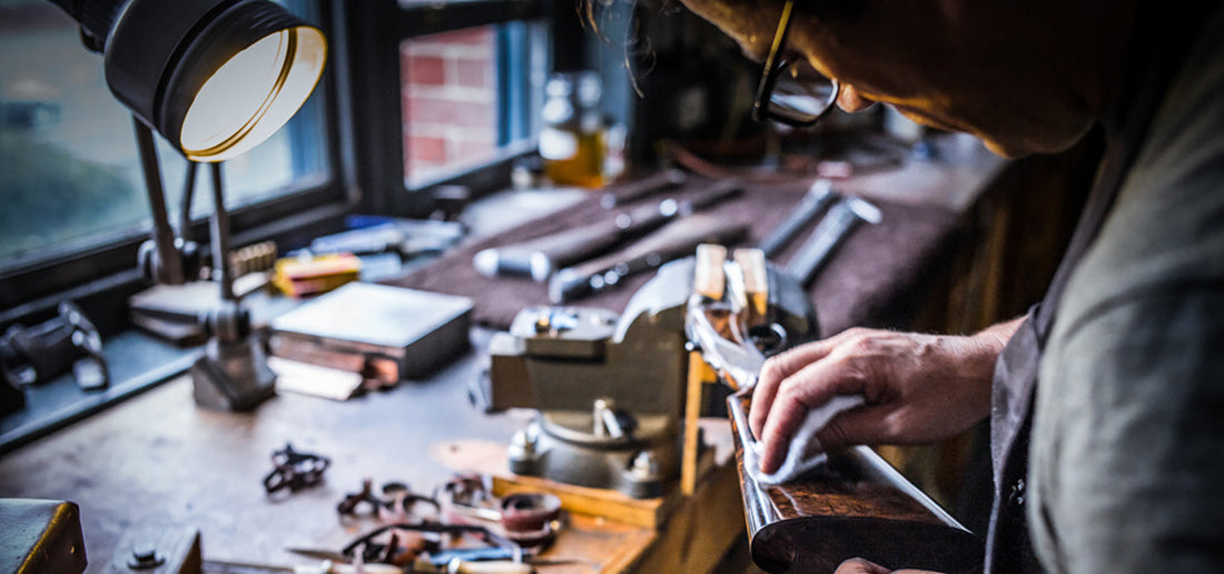 Gunmaker cleaning and inspecting a shotgun stock at a workbench under a lamp, showing careful firearm maintenance indoors.