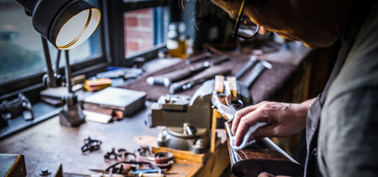 Gunmaker cleaning and inspecting a shotgun stock at a workbench under a lamp, showing careful firearm maintenance indoors.