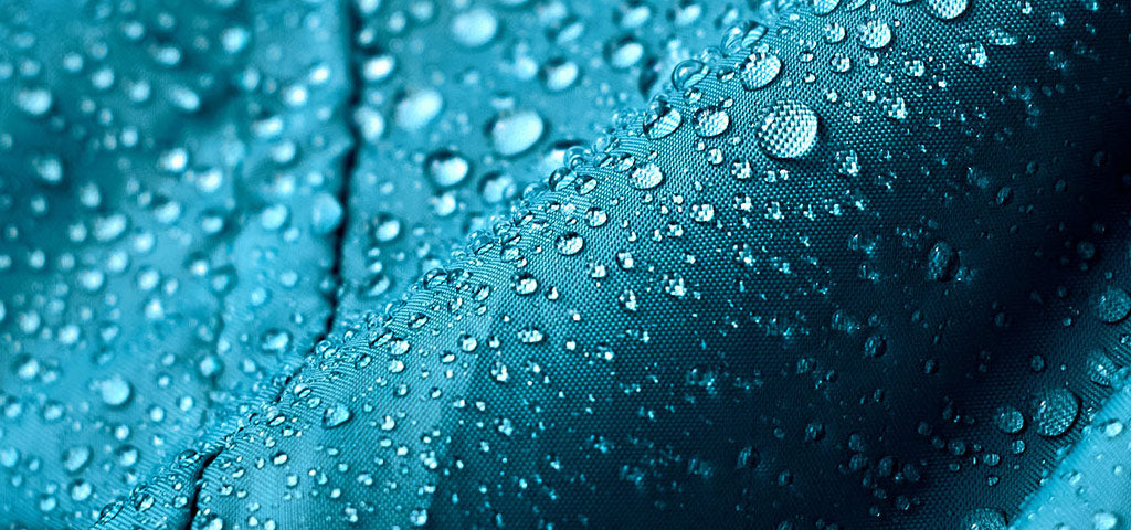 Close-up of a waterproof fabric in teal with water droplets demonstrating PFC-free water repellency.