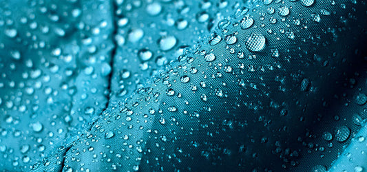 Close-up of a waterproof fabric in teal with water droplets demonstrating PFC-free water repellency.