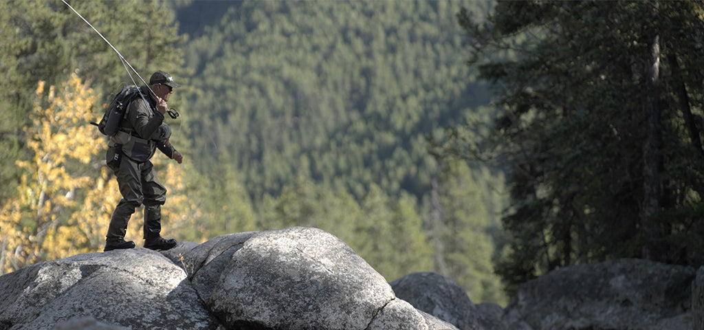 A man wearing Patagonia fishing gear, including Patagonia Swiftcurrent Waders, walks across large rocks with a fishing rod in hand, against a backdrop of a forested mountain slope.