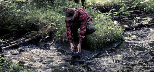 A man in a Sasta red plaid shirt collecting water from a forest stream.
