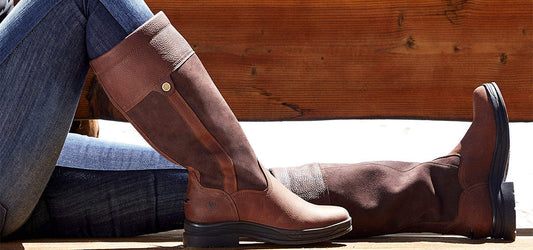 Woman reclining wearing blue jeans and Ariat brown leather riding boots against a wooden background.