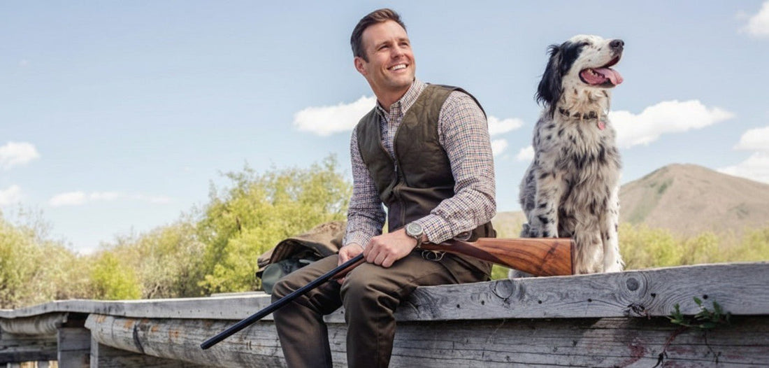 Man wearing Beretta shooting clothing sits on a wooden bridge with a gun and gundog.
