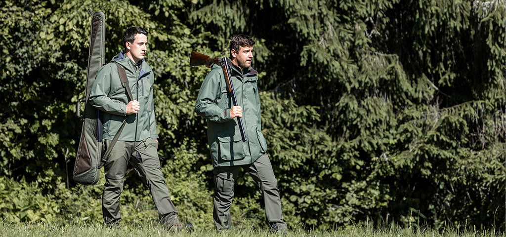 Two Men Walking Through Forest Wearing Shooting Jackets and Protective Clothing, Carrying Rifles in aGun Slip.