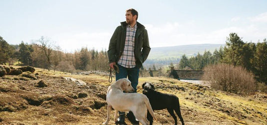 Man wearing Barbour outdoor clothing walking with two dogs in a scenic countryside. Ideal apparel for outdoor activities and dog walking in natural settings.