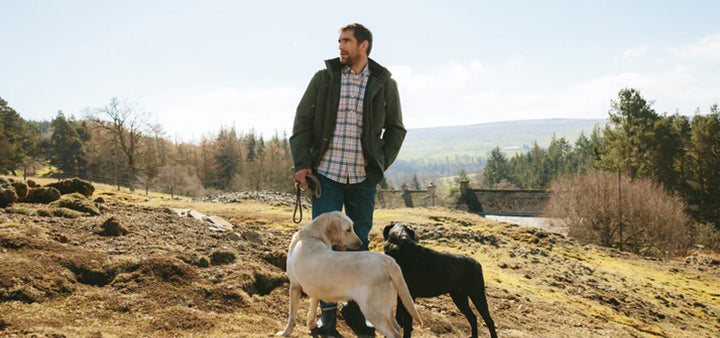 Man wearing Barbour outdoor clothing walking with two dogs in a scenic countryside. Ideal apparel for outdoor activities and dog walking in natural settings.