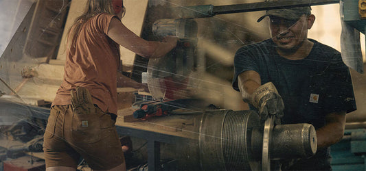 Two people working with tools and machinery, wearing Carhartt WIP workwear, in a dusty workshop environment.