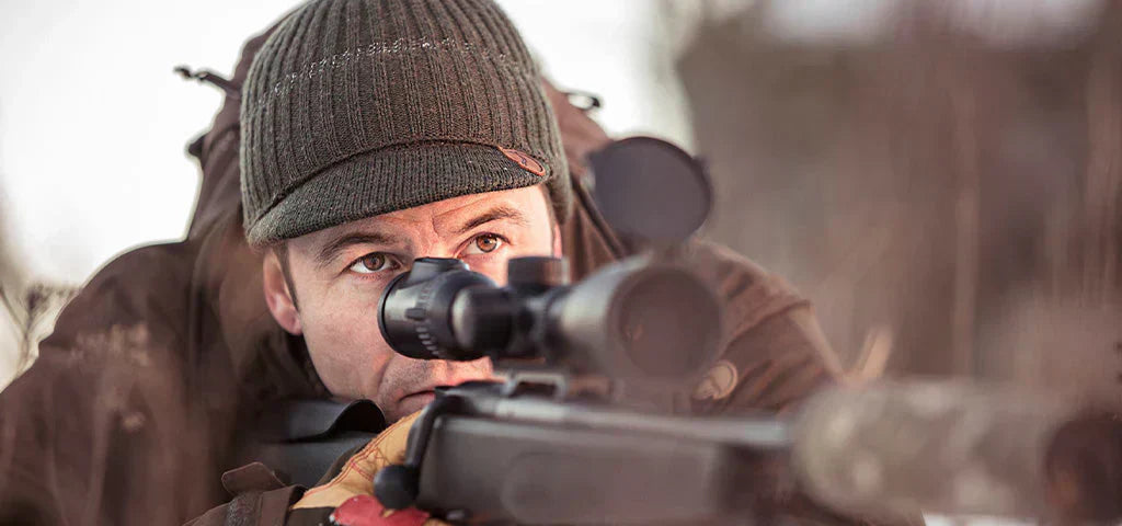 Man wearing Fjällräven shooting jacket and beanie aiming a rifle in winter field conditions, showing practical country shooting wear
