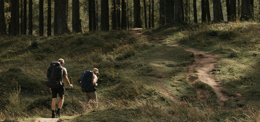 Two hikers with Fjallraven backpacks walking on a trail in the Peak District, surrounded by tall trees and grassy terrain.
