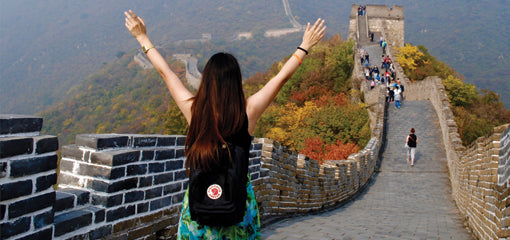 A woman with her arms raised in joy, wearing a Kanken backpack, stands on the Great Wall of China with autumn foliage in the distance.
