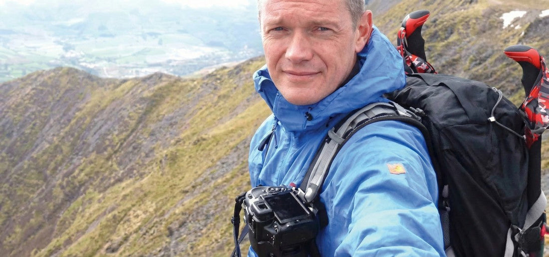 Phil Raisbeck wearing a blue Fjallraven High Coast Wind Jacket and carrying a black backpack while hiking in the mountains.