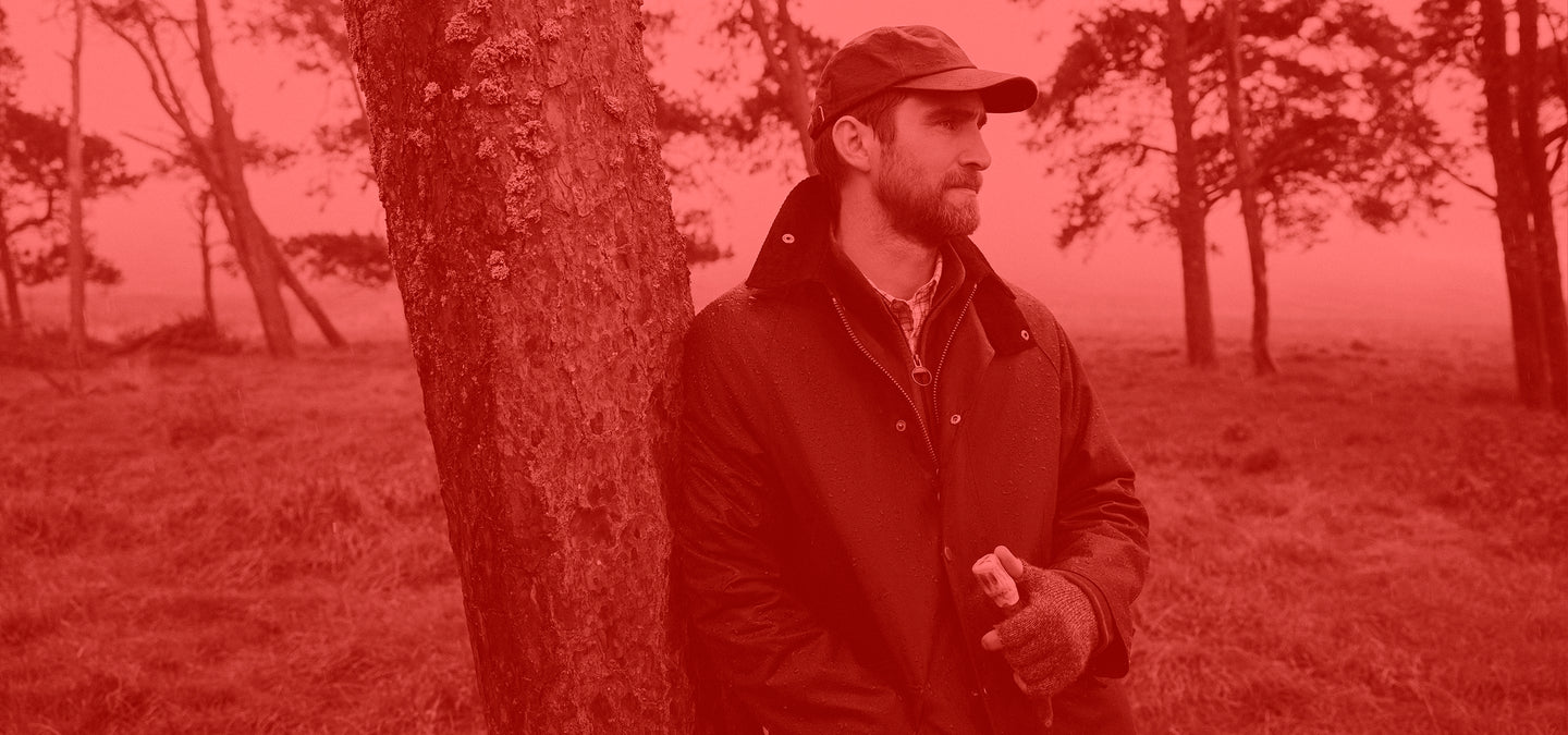 Man wearing Barbour Jacket abd Cap standing next to a tree in a field with a red filter