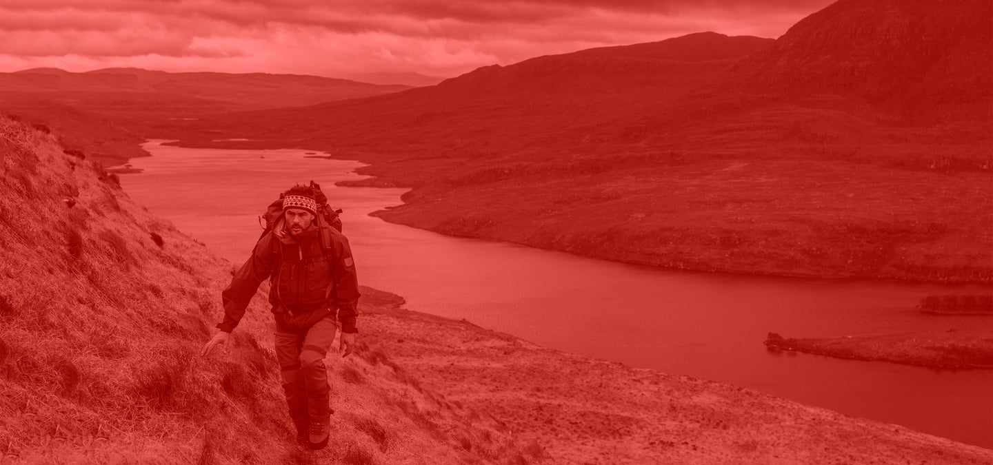 Man wearing Fjallraven clothing and backpack hiking along a path with a scenic view of a lake and mountains.
