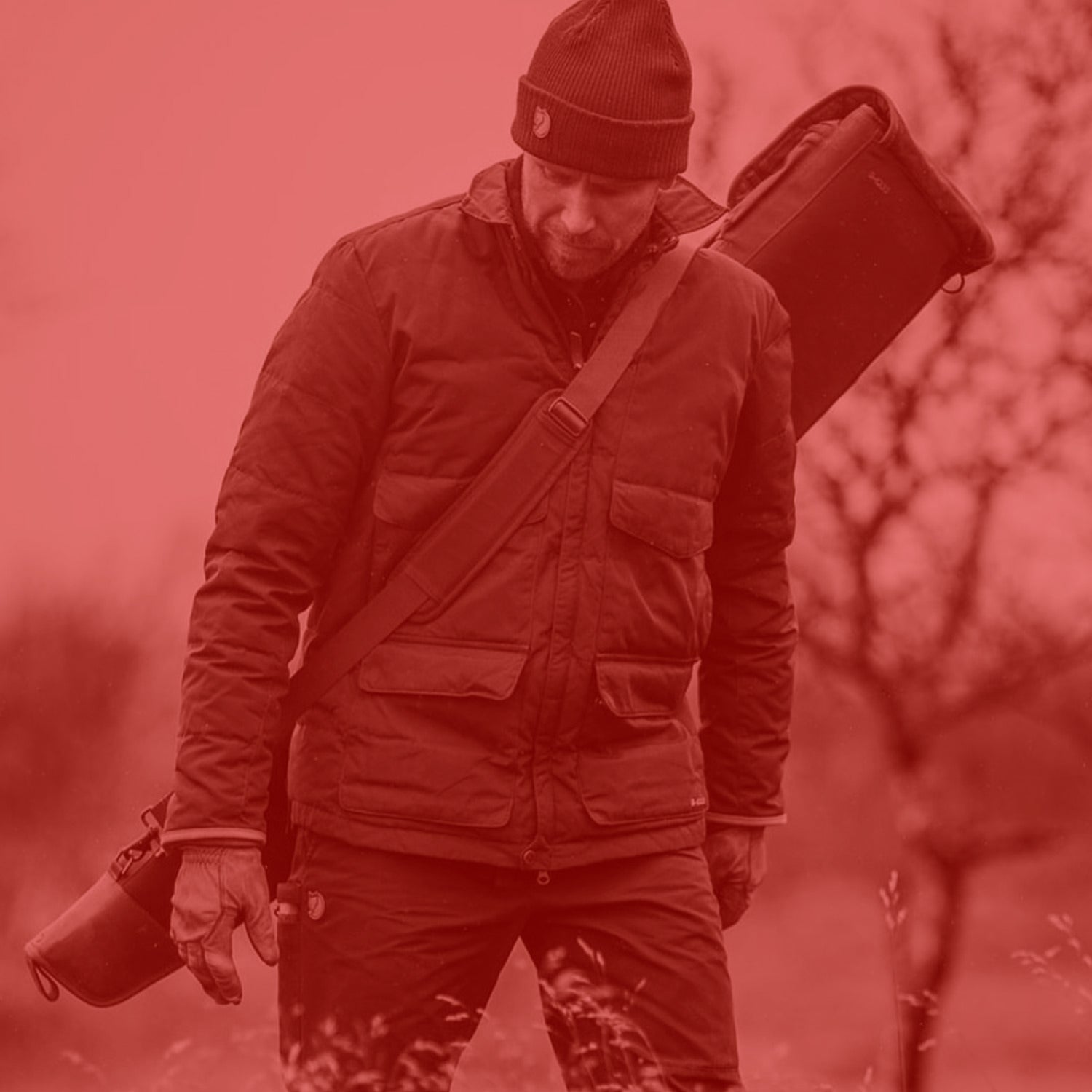 Man in outdoor gear carrying a gun case in a red-tinted forest setting