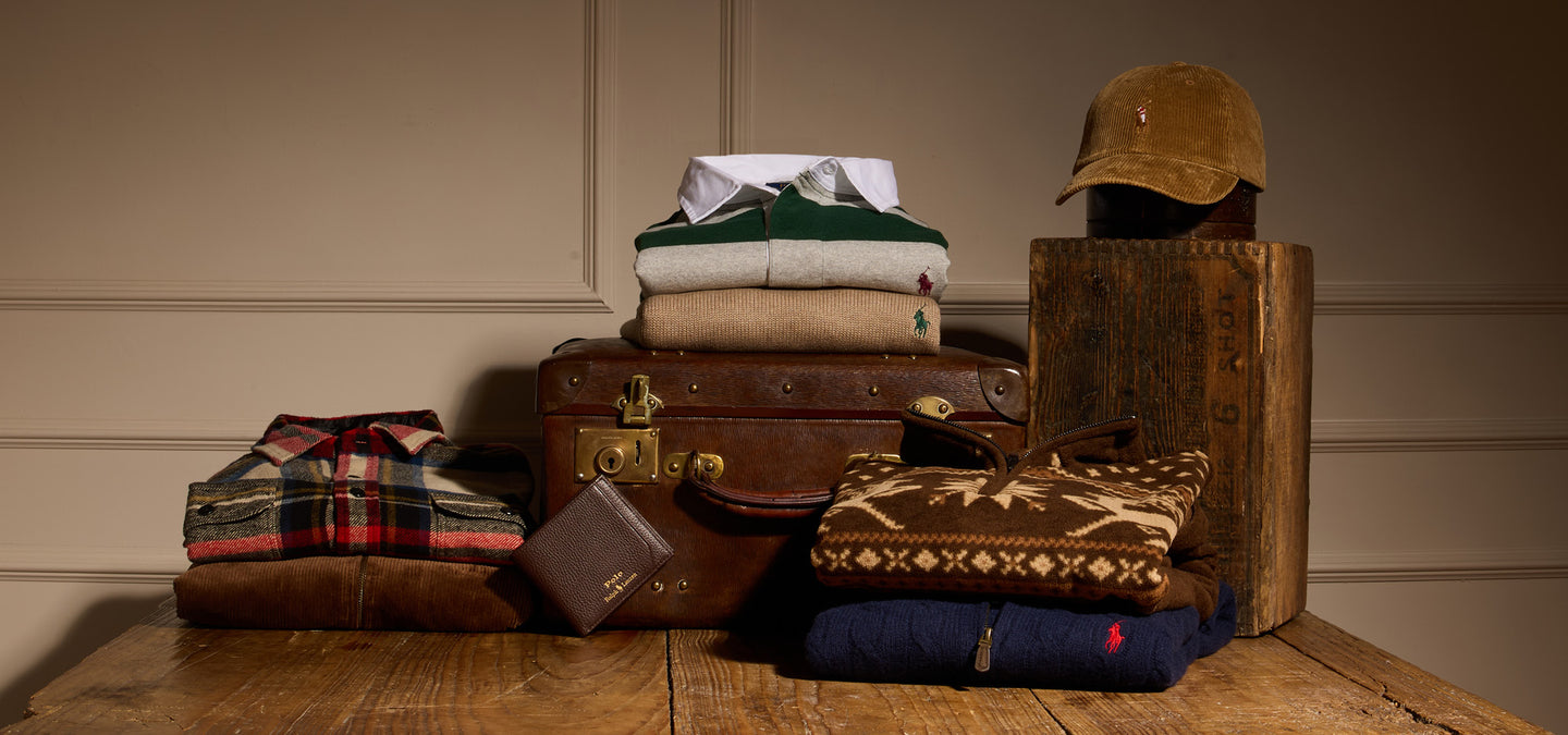 Stack of folded Polo Ralph Lauren clothes and a suitcase on a wooden surface with a neutral background.
