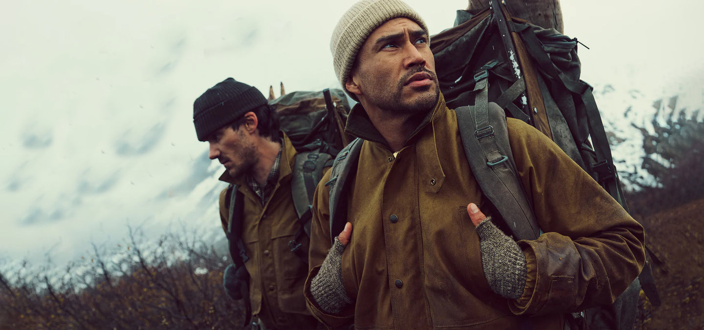 Two men wearing Filson jackets with backpacks walking through a forested area with a cloudy sky.