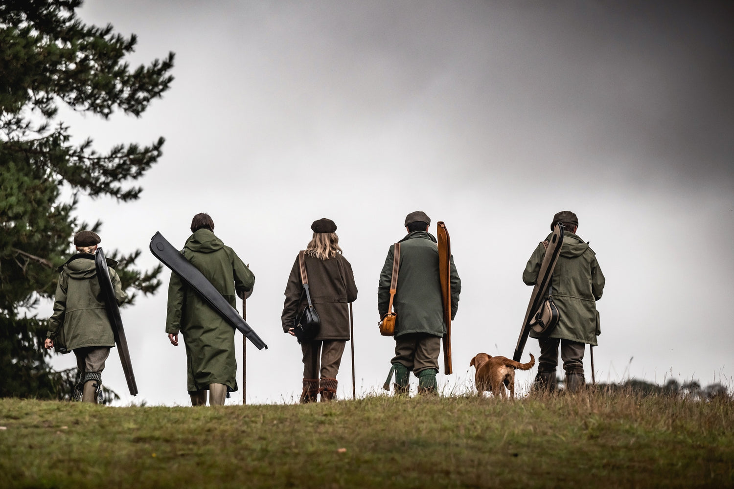 Five people in hunting gear with rifles on a grassy field, one dog visible.